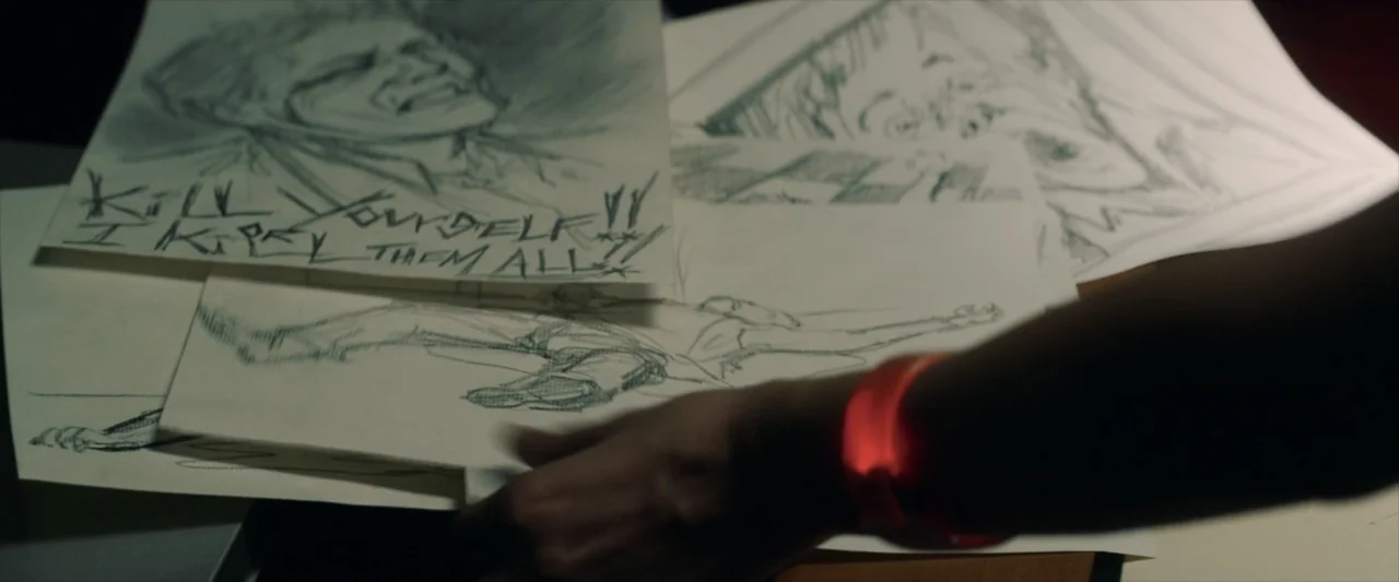 Tell Me How I Die (2016) movie still: closeup — A dark surface is covered with scattered hand-drawn sketches featuring disturbing text and figures.…