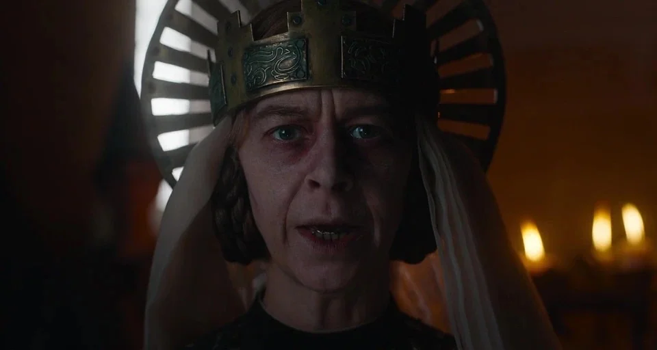 The Green Knight (2021) movie still: closeup — A dimly lit stone environment contains a woman wearing a distinctive metallic crown. She faces the…
