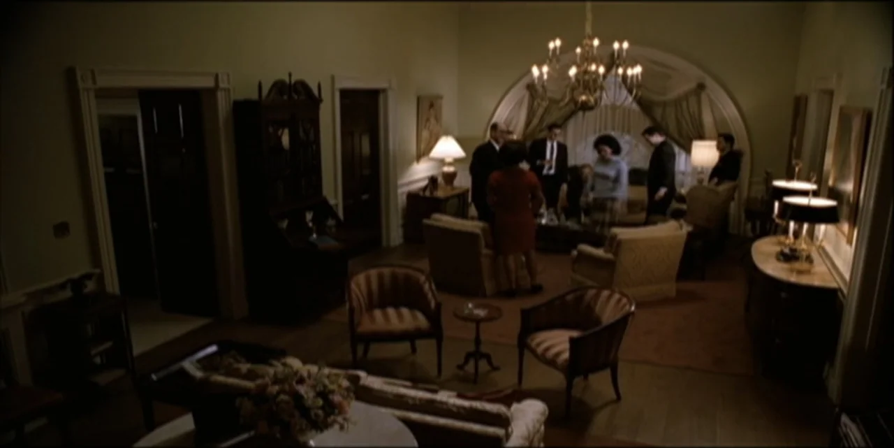 Path to War (2003) movie still: wide — A spacious, dimly lit formal sitting room features mid-century furniture and a large chandelier. Several…