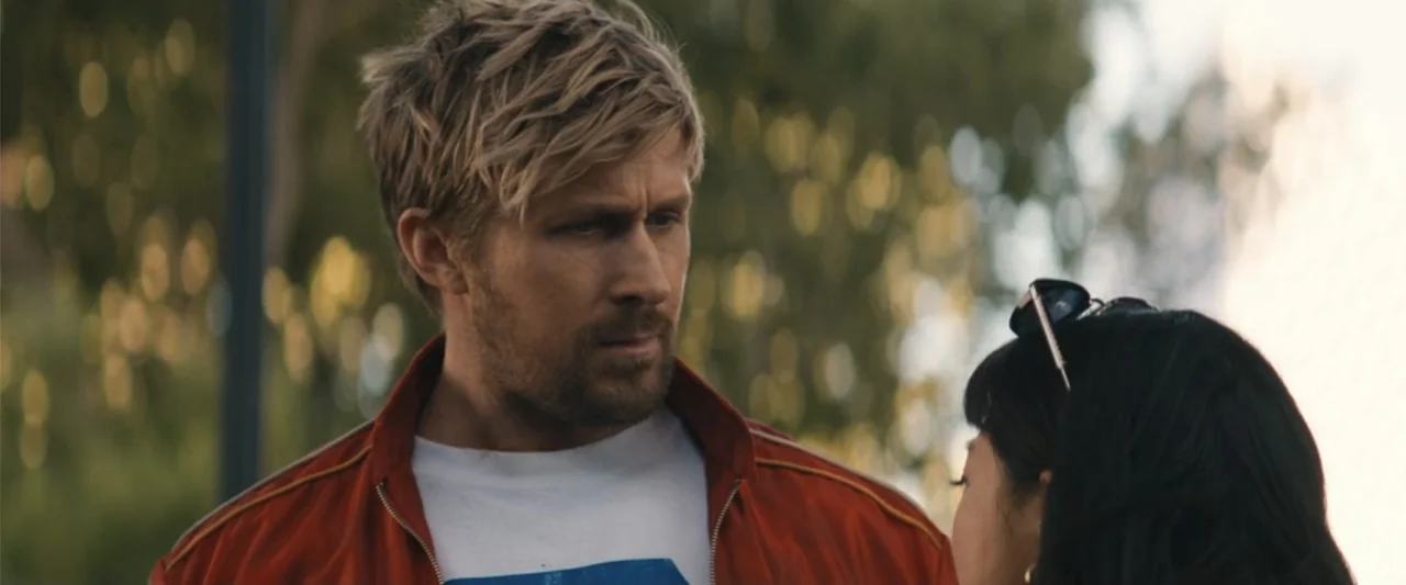 The Fall Guy (2024) movie still: medium — A man and woman stand in an outdoor park area with blurred green foliage. The man in a red jacket gazes…