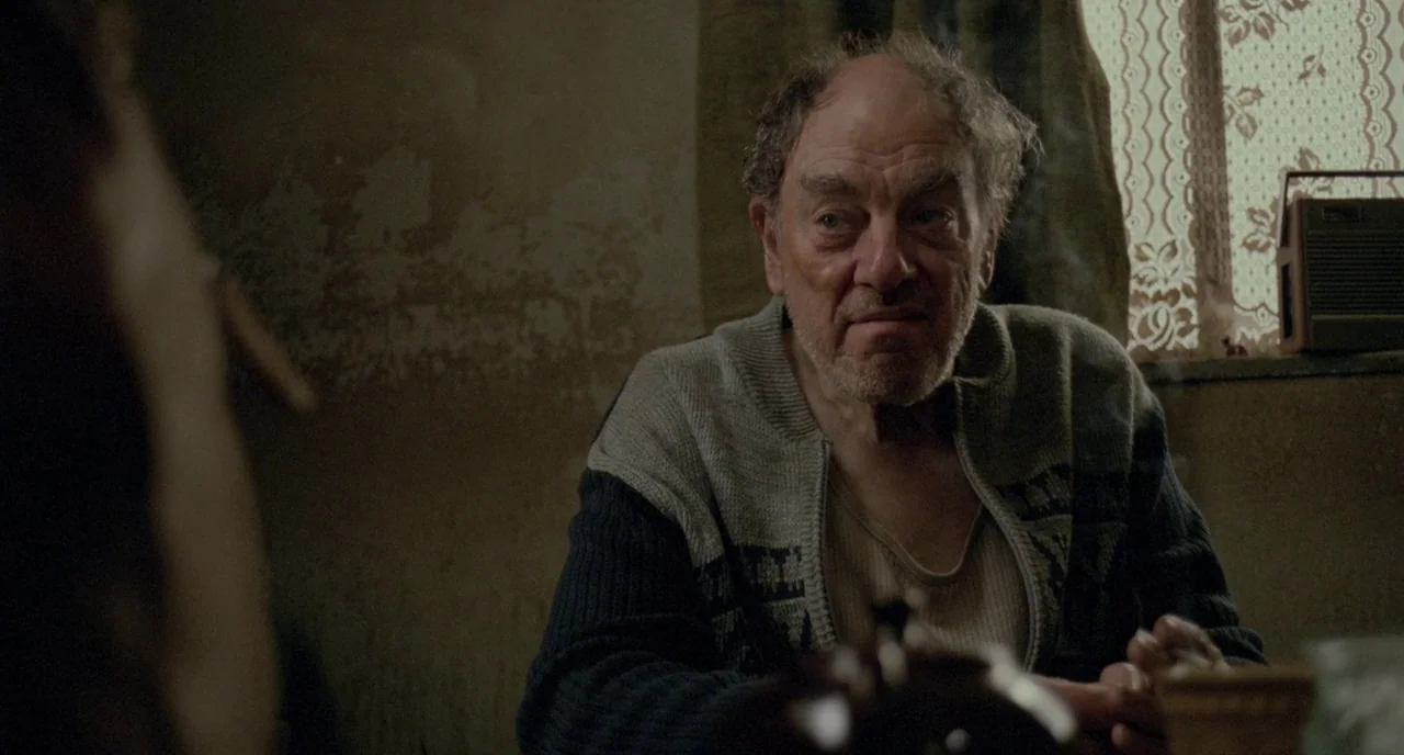 Possum (2018) movie still: medium — An aged man sits inside a room with plastered walls and a patterned window curtain. He holds a lit cigarette…