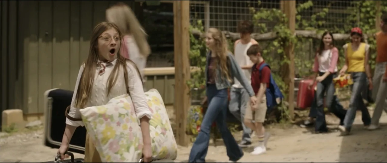Summer Camp (2024) movie still: medium — A group of teenagers carries suitcases along a dirt path at a summer camp. One girl in the foreground walks…
