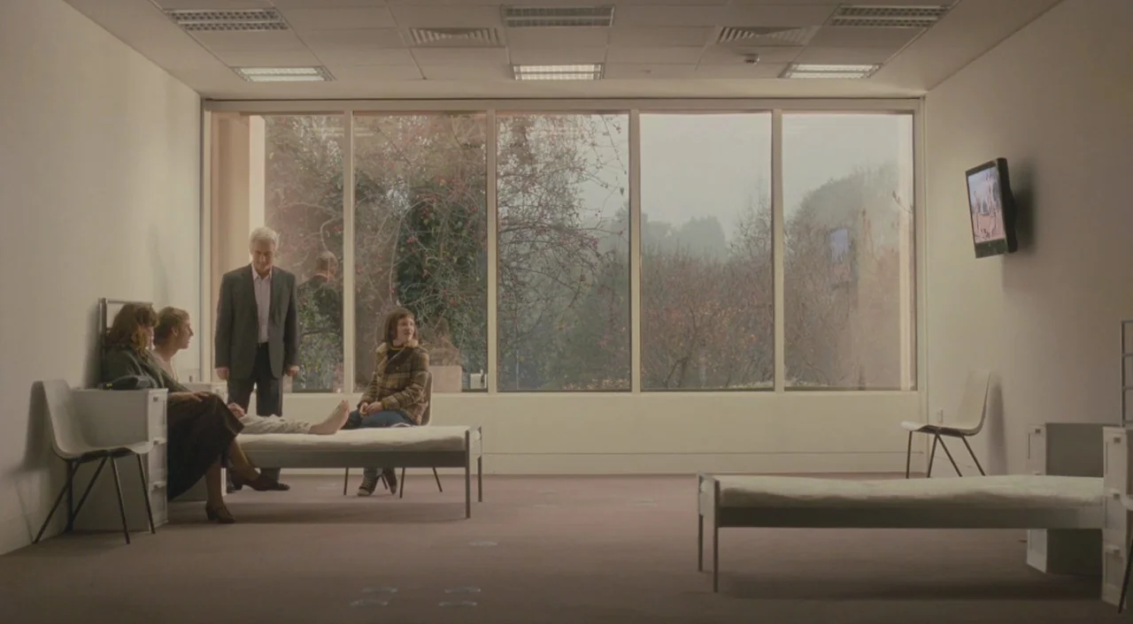 Broken (2012) movie still: wide — A sterile room with large windows showing bare trees outside in gray weather. Four people sit or stand around two…