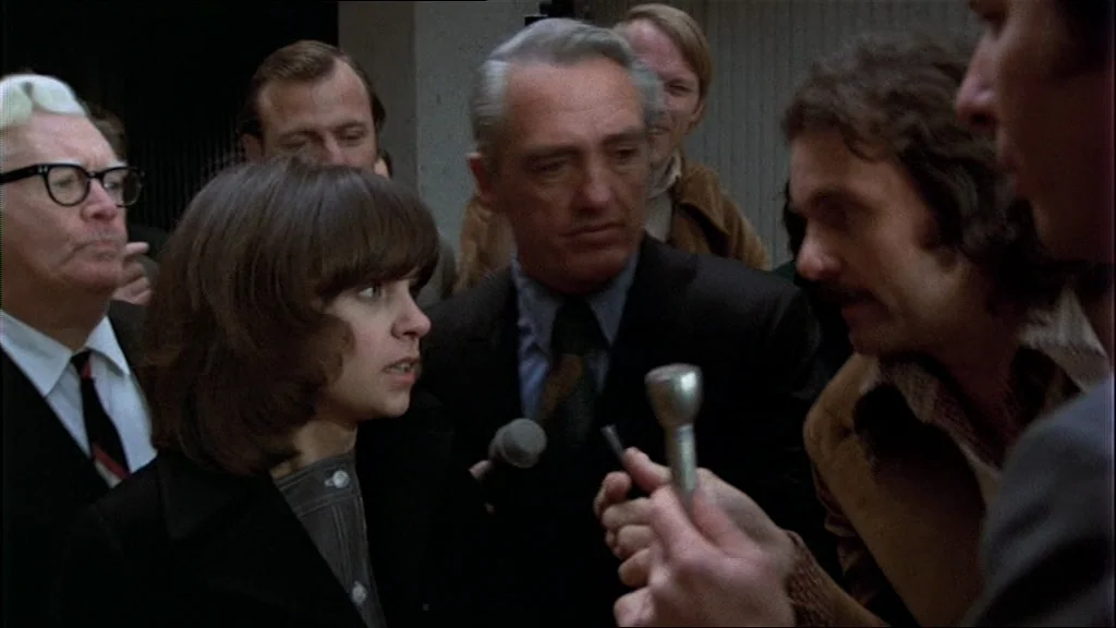 The Conversation (1974) movie still: medium — A crowd of people gathers on a city street for a press encounter. A woman in a dark coat speaks into…