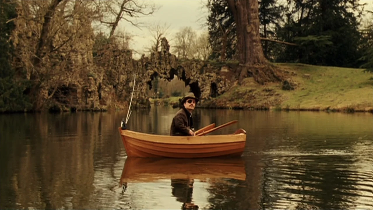 Penelope (2007) movie still: wide — A calm, reflective pond is surrounded by manicured grassy banks and mature leafless trees. A man wearing an…