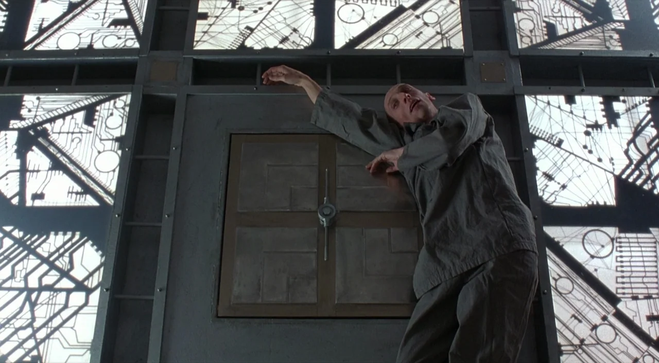 Cube (1997) movie still: fullbody — A metallic wall features backlit translucent panels etched with intricate, glowing black circuit board diagrams.…