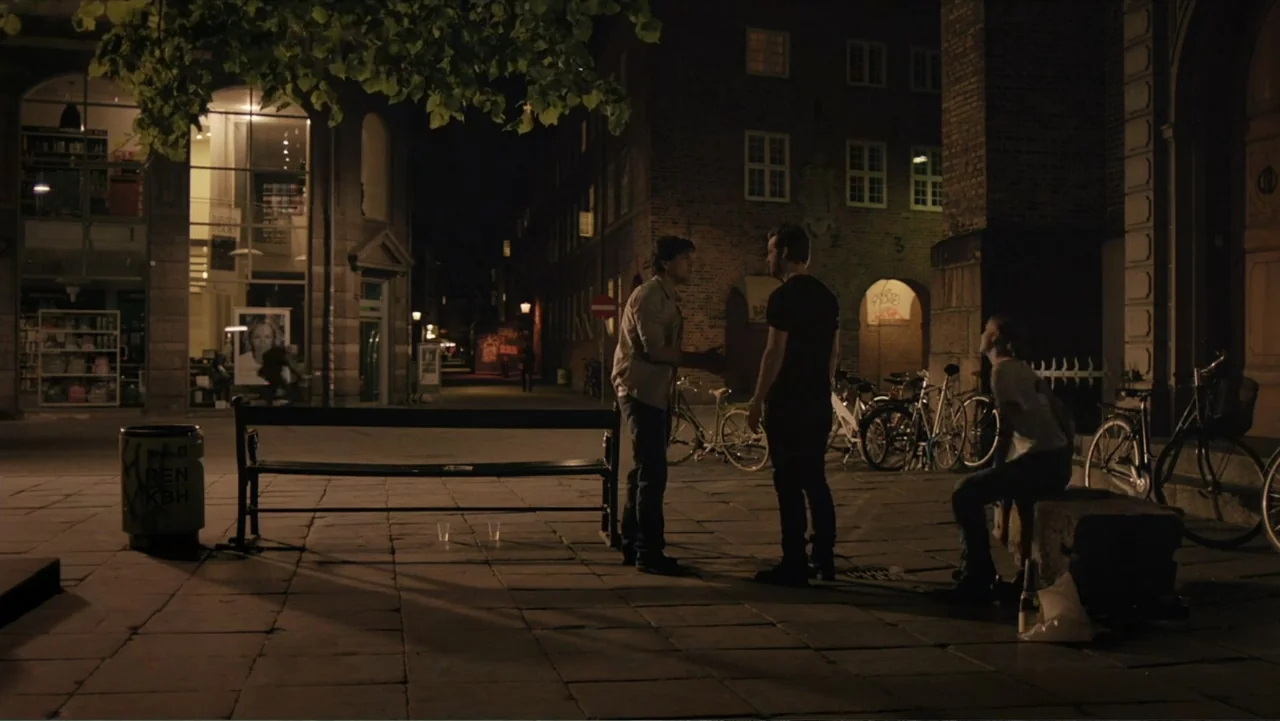Copenhagen (2002) movie still: wide — A stone paved city square at night features a wooden bench, bicycles, and brick buildings. Three young people…