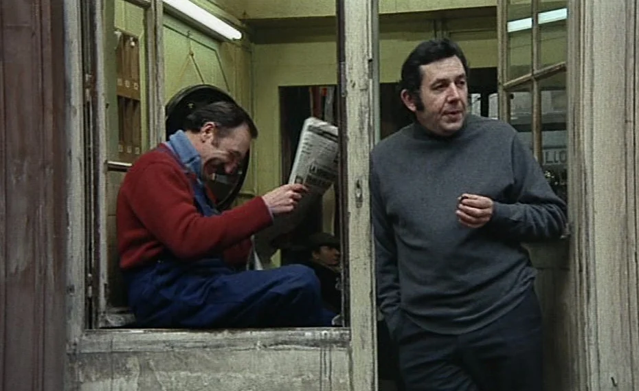 Bed & Board (1970) movie still: medium — A cramped, beige-walled concierge booth features a large window frame and wooden door frame. Two…