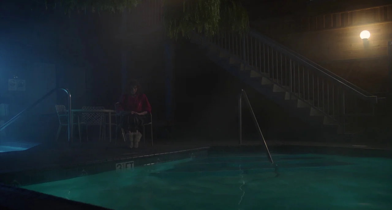 An Evening With Beverly Luff Linn (2018) movie still: wide — A dark outdoor swimming pool area at night with stairs leading up to rooms. A lone…
