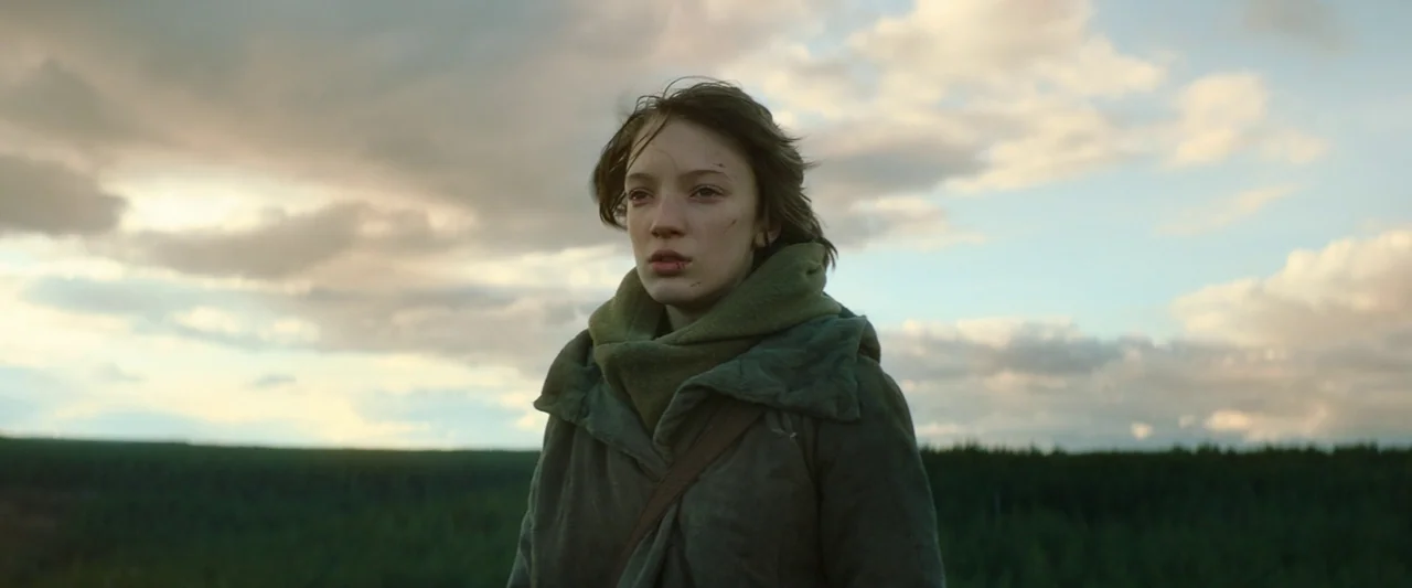 Vesper (2022) movie still: medium — An open field under a cloudy, overcast sky during the late afternoon. A teenage girl in a green jacket stands in…