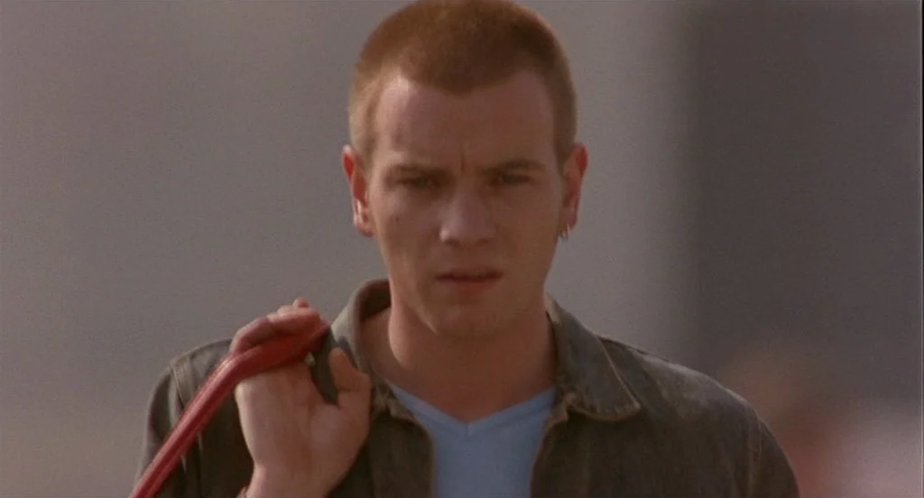 Trainspotting (1996) movie still: medium — A man with a shaved head stands in an indeterminate outdoor setting. He faces the camera, carrying a red…