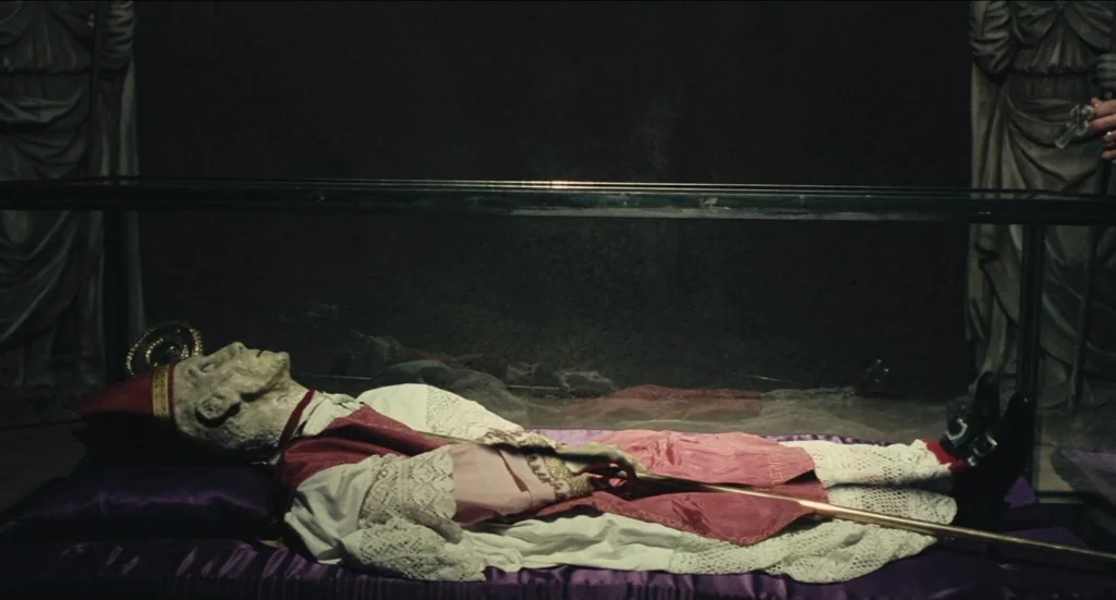 Todo Modo (1976) movie still: wide — A mummified figure dressed in religious regalia lies inside a glass-topped casket. The setting is a dark,…