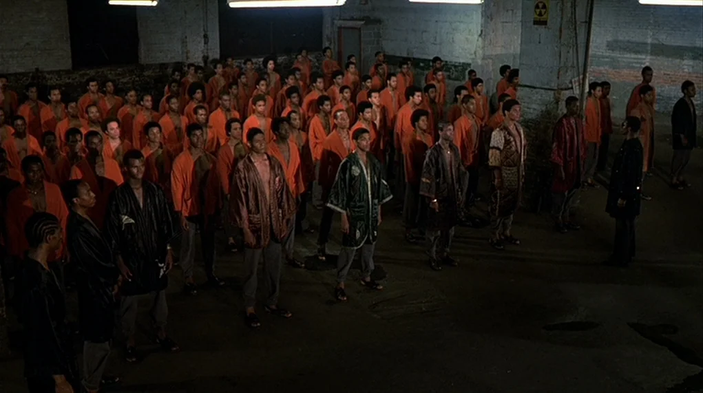 The Warriors (1979) movie still: wide — A vast, dark industrial space contains a large crowd of men wearing matching robes. The men stand in ordered…