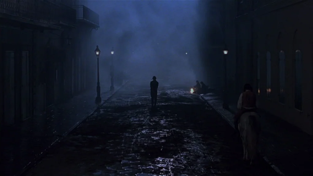 Interview With The Vampire movie still: wide — A dark, misty cobblestone road at night is illuminated by dim streetlamps. A lone man walks ahead…