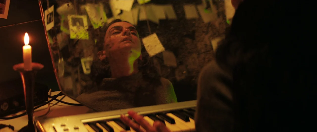In The Earth (2021) movie still: medium — A dim room contains a keyboard and a mirror adorned with numerous white paper notes. The woman looks…