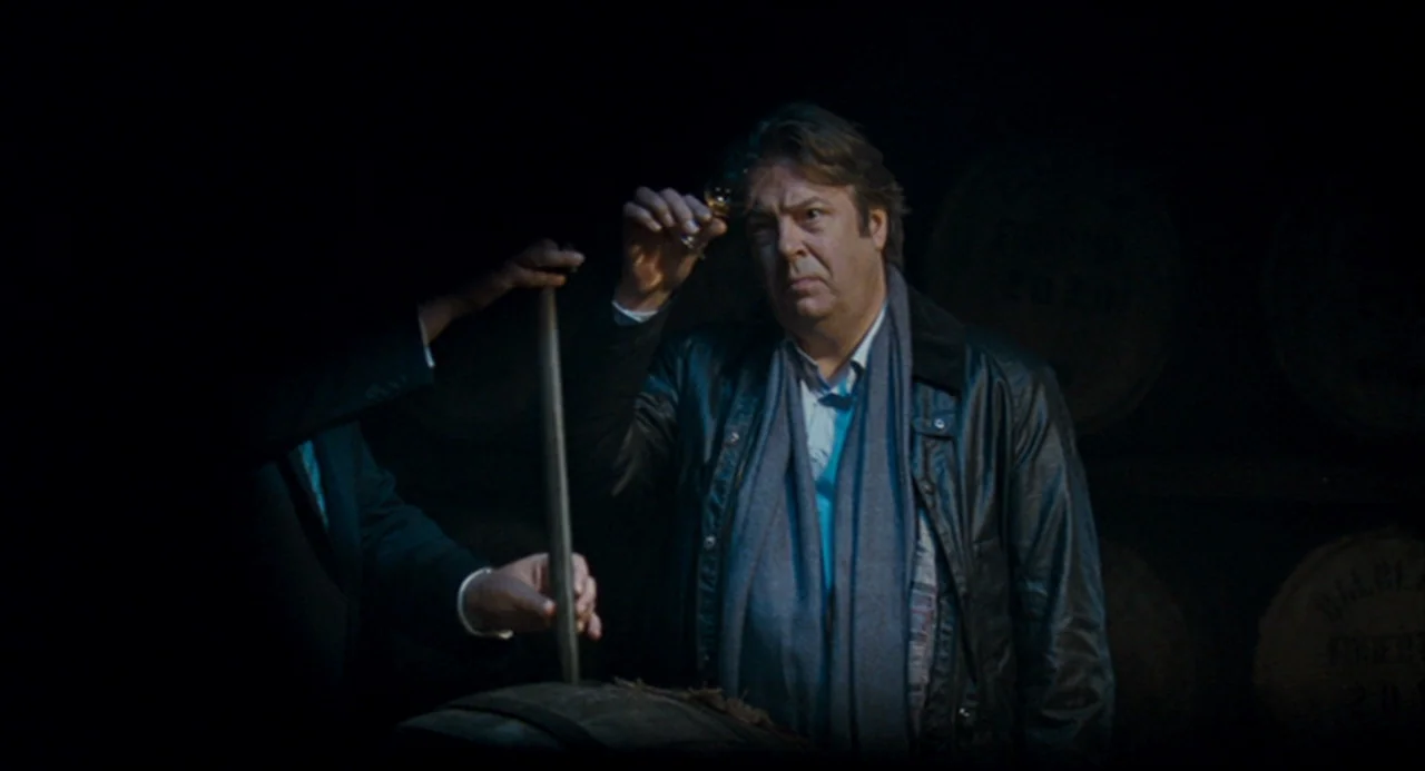 The Angel’s Share (2012) movie still: medium — A dark warehouse filled with wooden casks arranged in rows along the walls. A man holds a small…