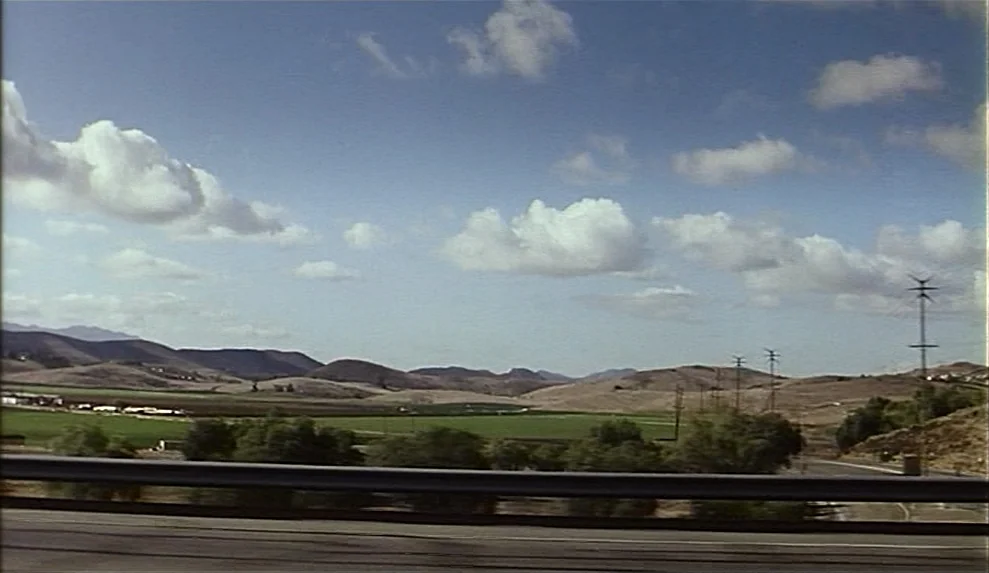 Safe movie still: establishing — A view from a highway showing rolling hills and green fields under a sky. A metal guardrail runs horizontally…