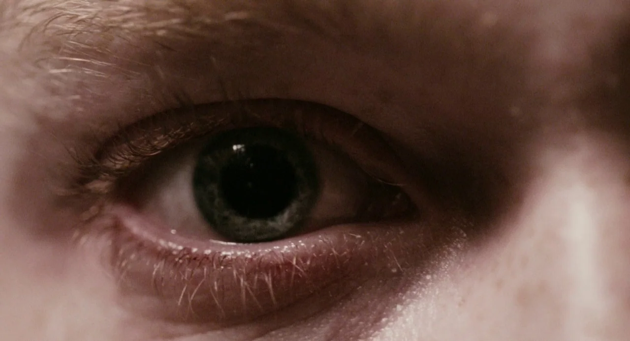 Antiviral (2012) movie still: closeup — A extreme close-up of a human eye with a dark, textured iris. Fine skin details and individual hair…