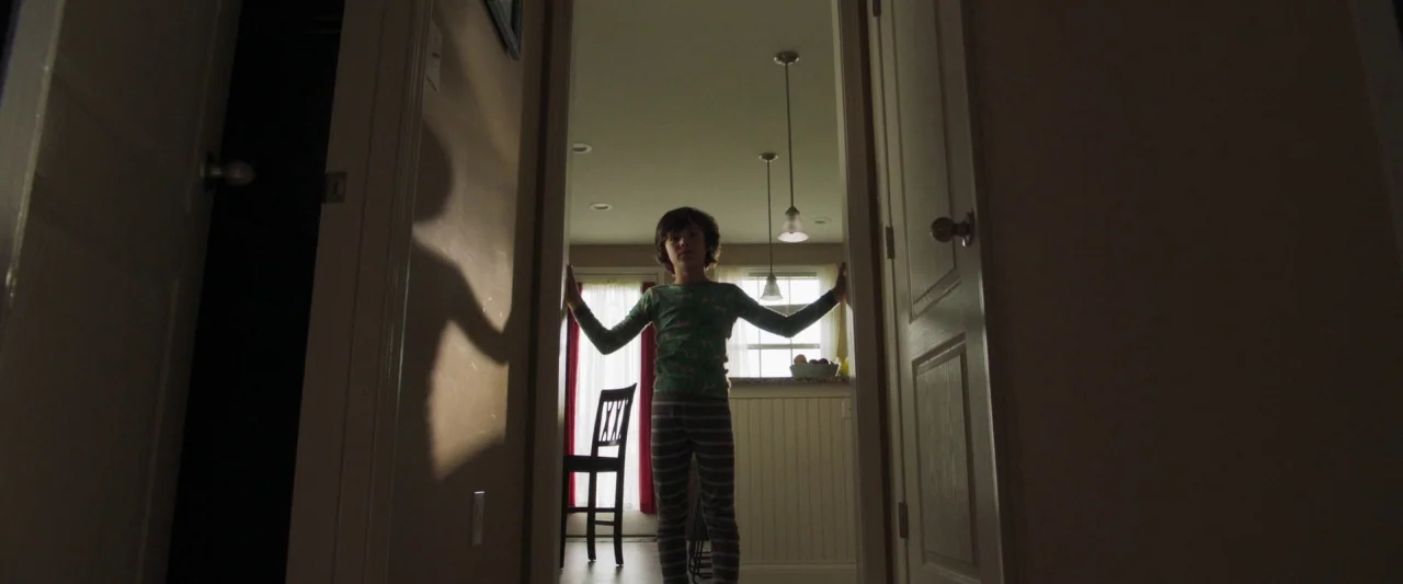 Mom and Dad (2017) movie still: fullbody — A dark hallway leads into a bright room with white walls and a window. A young boy stands in the doorway…