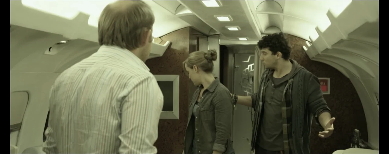 Turbulence (2025) movie still: medium — The interior of an airplane cabin features wood-paneled walls and recessed overhead lights. A woman stands…