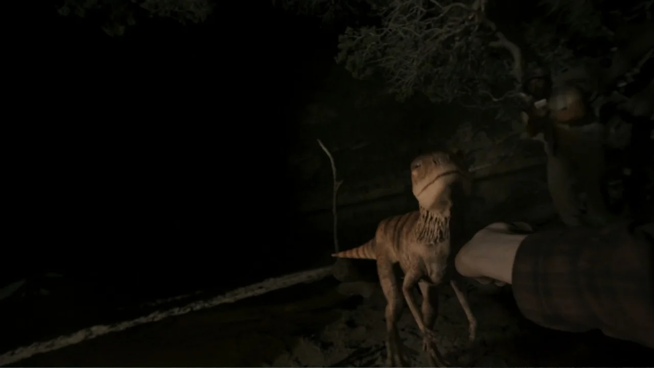 The Dinosaur Project (2012) movie still: medium — A dark outdoor space at night shows a small dinosaur standing near trees. A person reaches out a…
