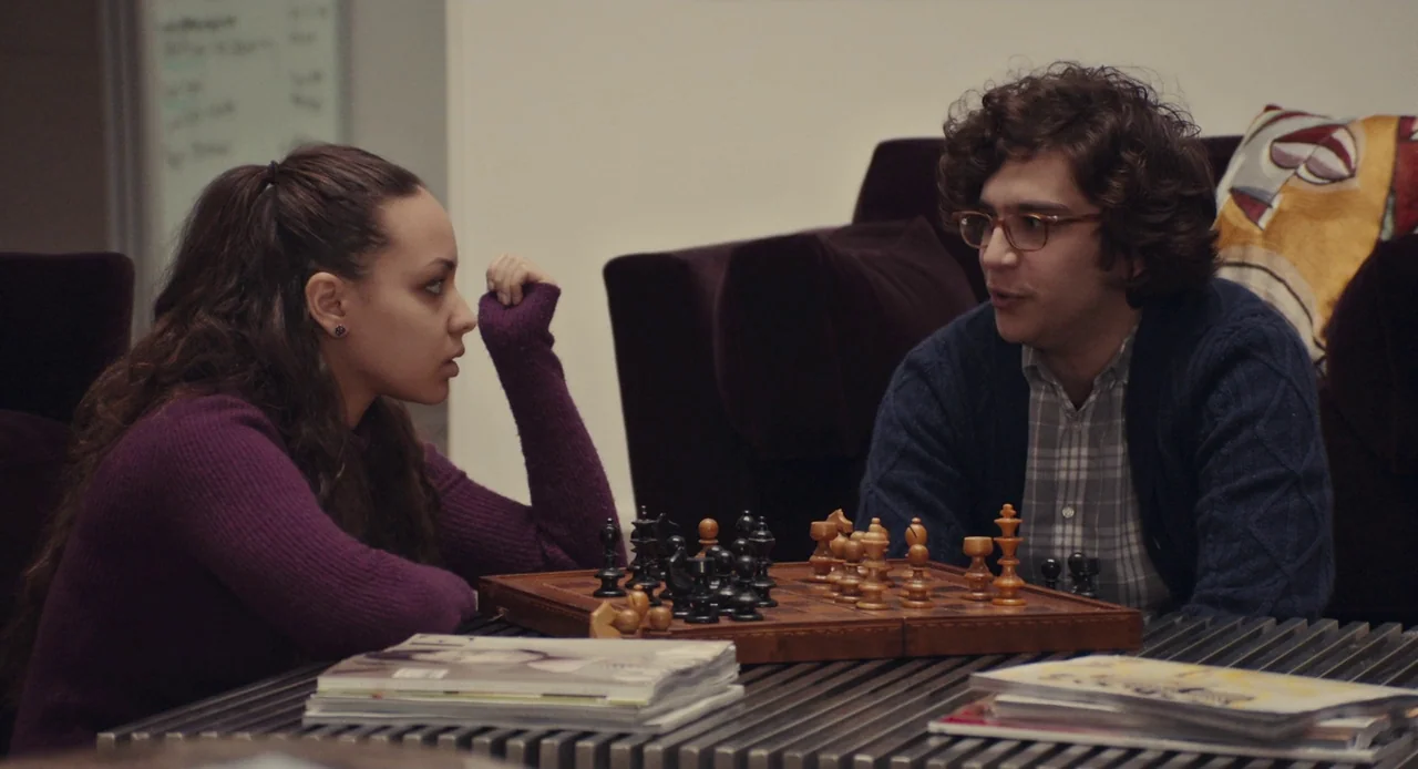 Mistress America (2015) movie still: medium — A quiet common room area with deep purple armchairs and a metallic coffee table. A young man and woman…
