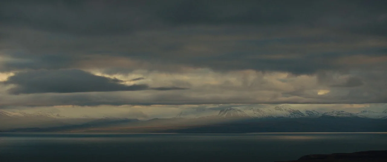 Macbeth (2015) movie still: establishing — A vast lake stretches beneath a mountain range dusted with patches of snow. Heavy, dark clouds blanket…