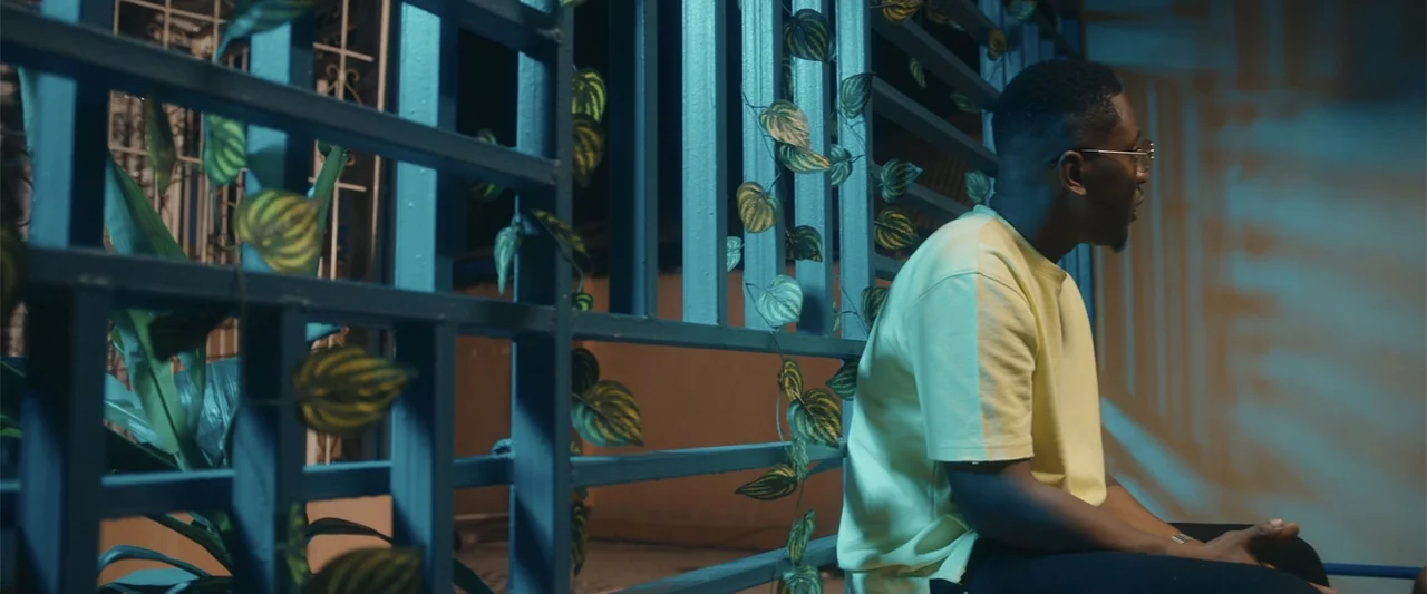 Big Love (2012) movie still: medium — A man sits against a metal gate with artificial leaves attached during the night. The man wearing a yellow…