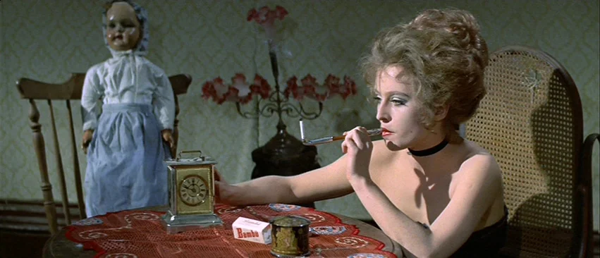 Whity (1971) movie still: medium — A woman sits at a round table featuring a clock and smoking materials. An antique porcelain doll is positioned…