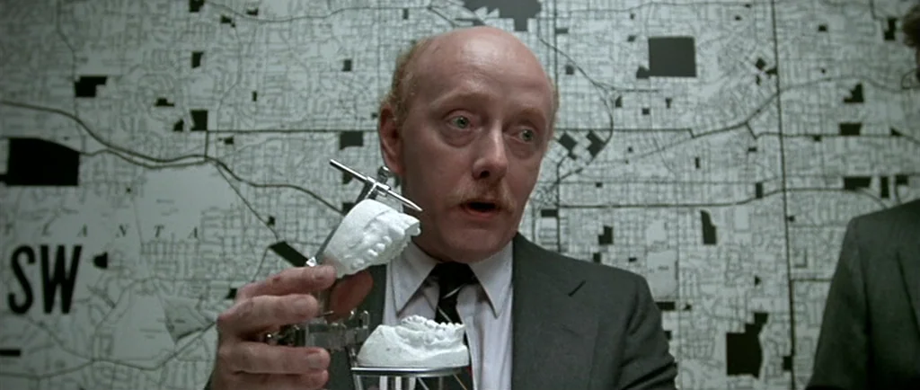 Manhunter (1986) movie still: medium — An interior space features a large black-and-white grid map on the wall. A man in a suit displays a white…