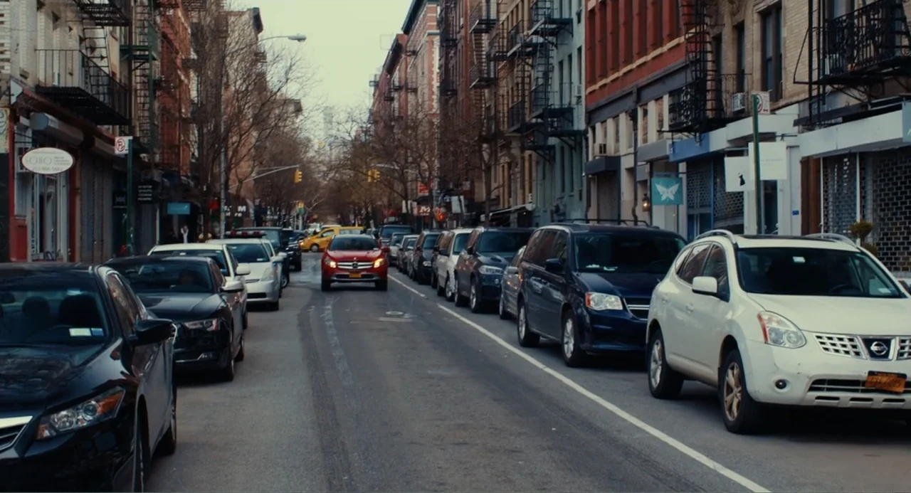An Imperfect Murder (2017) movie still: wide — A narrow New York City street features multi-story brick buildings with fire escapes. Multiple cars…