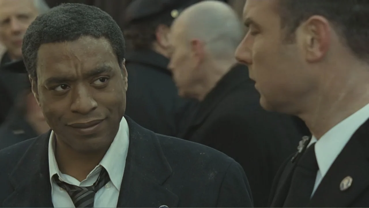 Salt (2010) movie still: medium — A group of people dressed in dark formal attire stand outdoors during a funeral service. One man in a black suit…