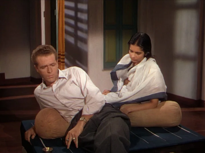 The River (1951) movie still: medium — A quiet indoor space with traditional cushions resting on a dark blue platform base. A man and a woman sit…