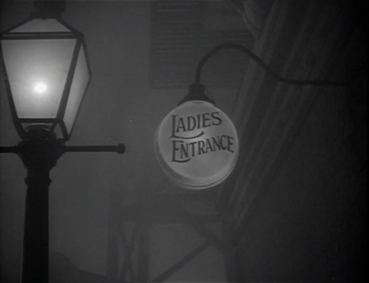 Anna Christie (1930) movie still: wide — A dark, foggy exterior features a streetlamp and a sign reading Ladies Entrance. The sign glows against the…
