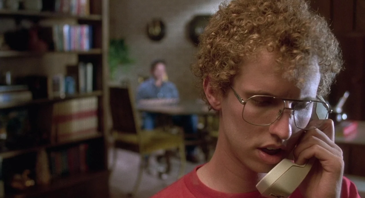 Napoleon Dynamite (2004) movie still: closeup — A living room interior features a bookshelf to the left and a dining area blurred in the background.…