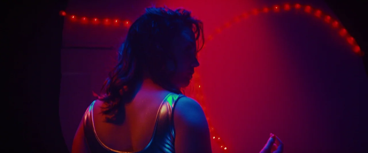 Revealer (2022) movie still: medium — A dark space defined by vibrant magenta light and curved lines of red LEDs. A woman in a shiny metallic top…
