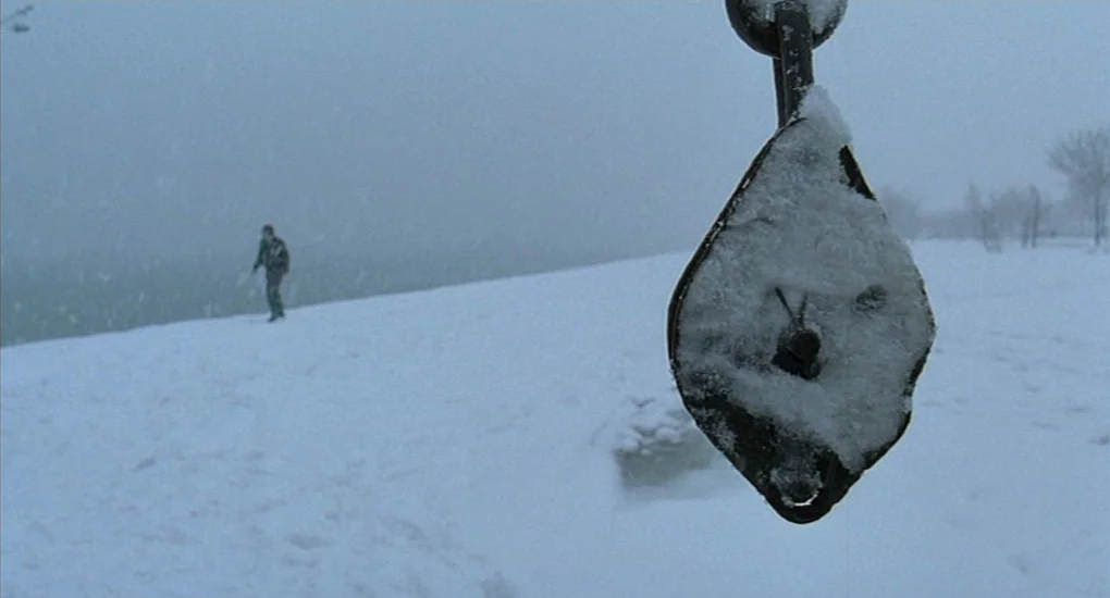 Uzak (2002) movie still: A hauntingly still shot featuring a snow-laden, rusted mechanical object in sharp focus, while a solitary figure wanders…