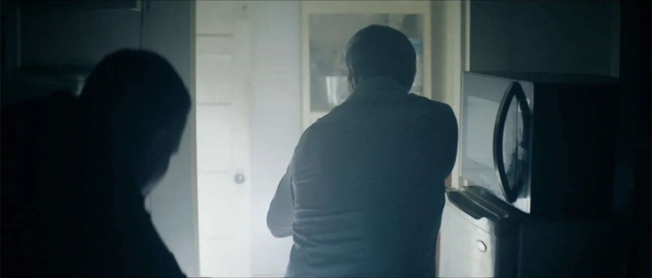 Crescent City (2024) movie still: medium — A dimly lit room features a microwave placed atop a refrigerator near a bright doorway. Two people stand…