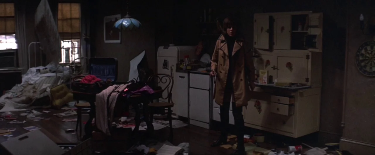 Klute (1971) movie still: fullbody — A dark apartment room is filled with scattered papers and luggage on the floor. A young woman in a trench coat…