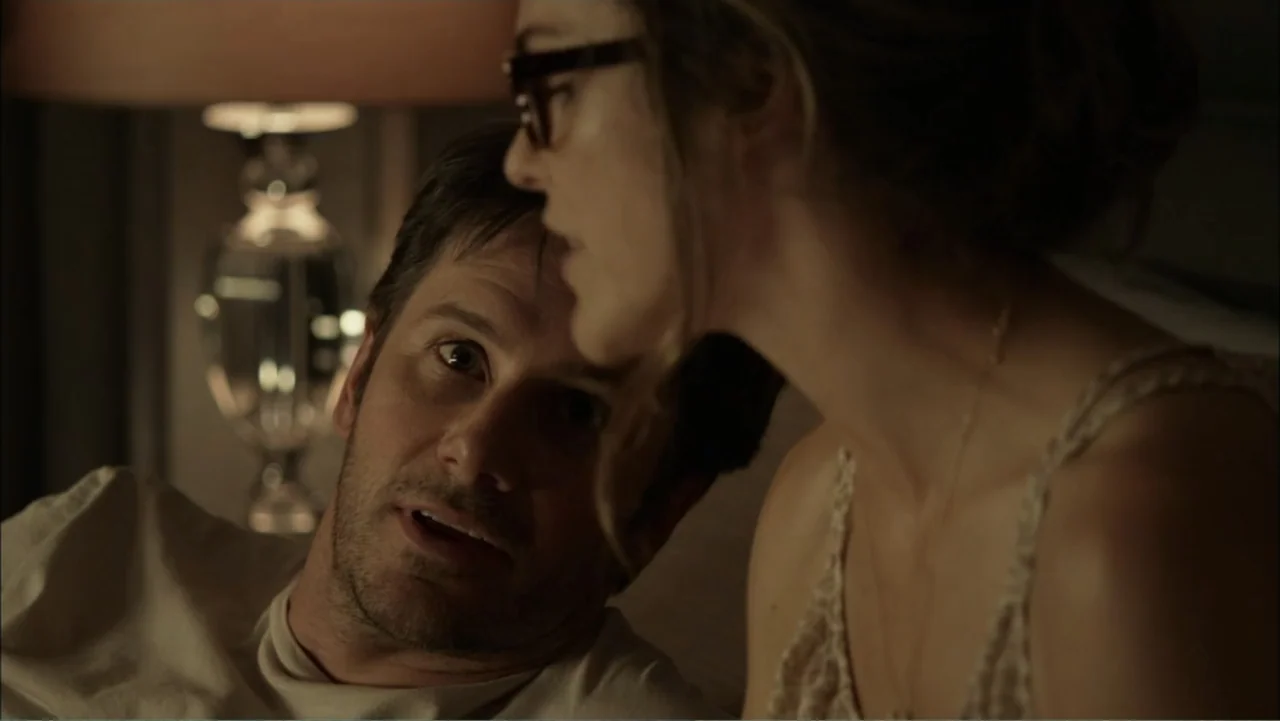 Dark Skies (2013) movie still: medium — A dark bedroom with a lit lamp visible in the background out of focus. A man lies in bed while a woman faces…