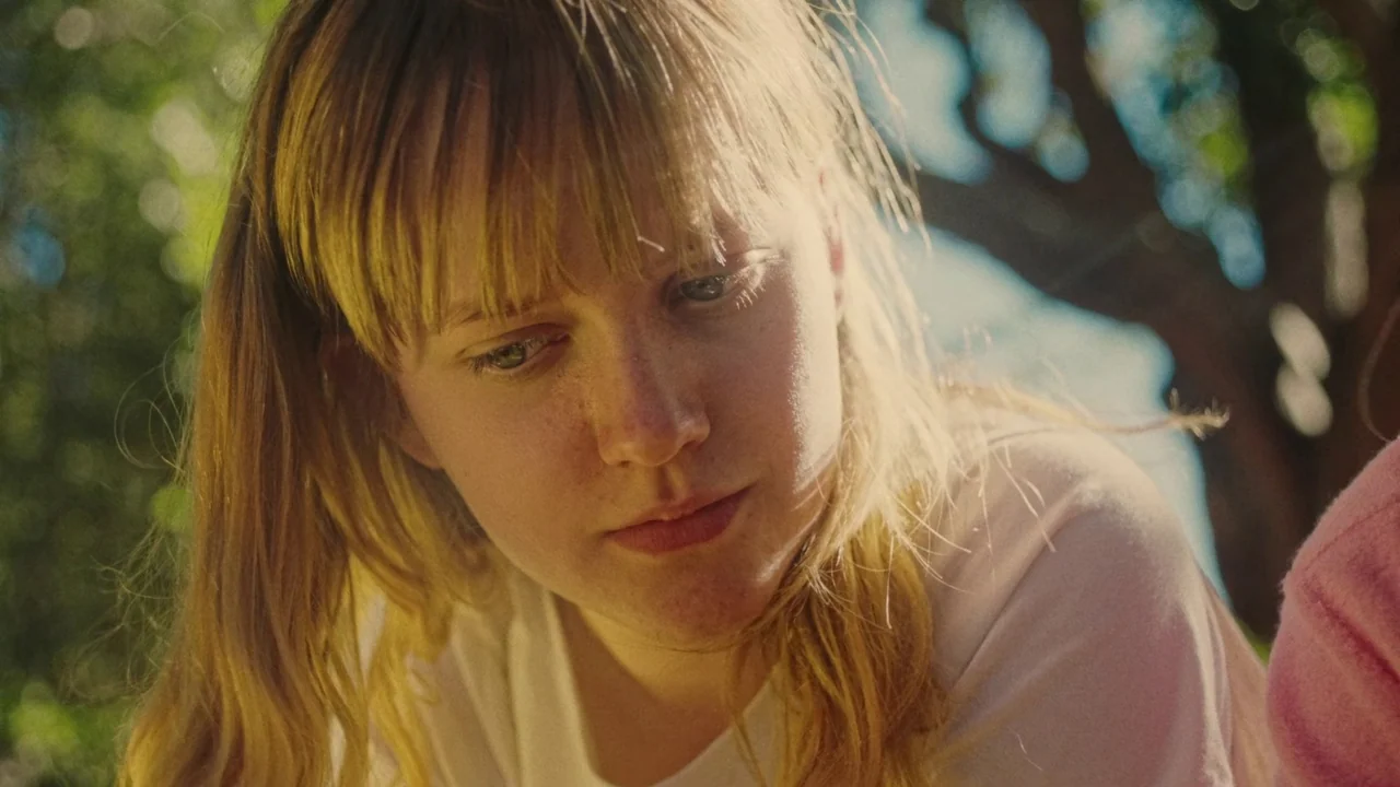 My First Summer (2020) movie still: closeup — An outdoor setting with blurred green foliage visible in the background under bright sunlight. A young…