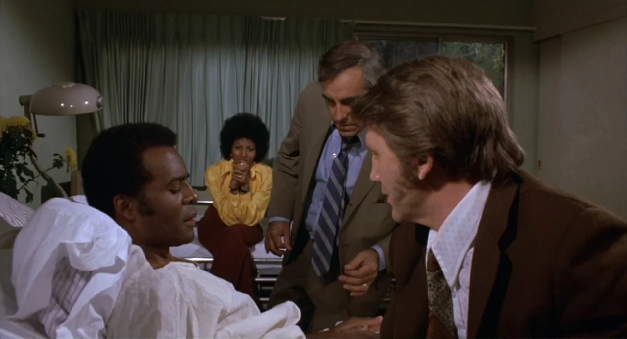 Foxy Brown (1974) movie still: medium — A hospital room features green curtains and a bed with three people gathered around. Two men in blazers…