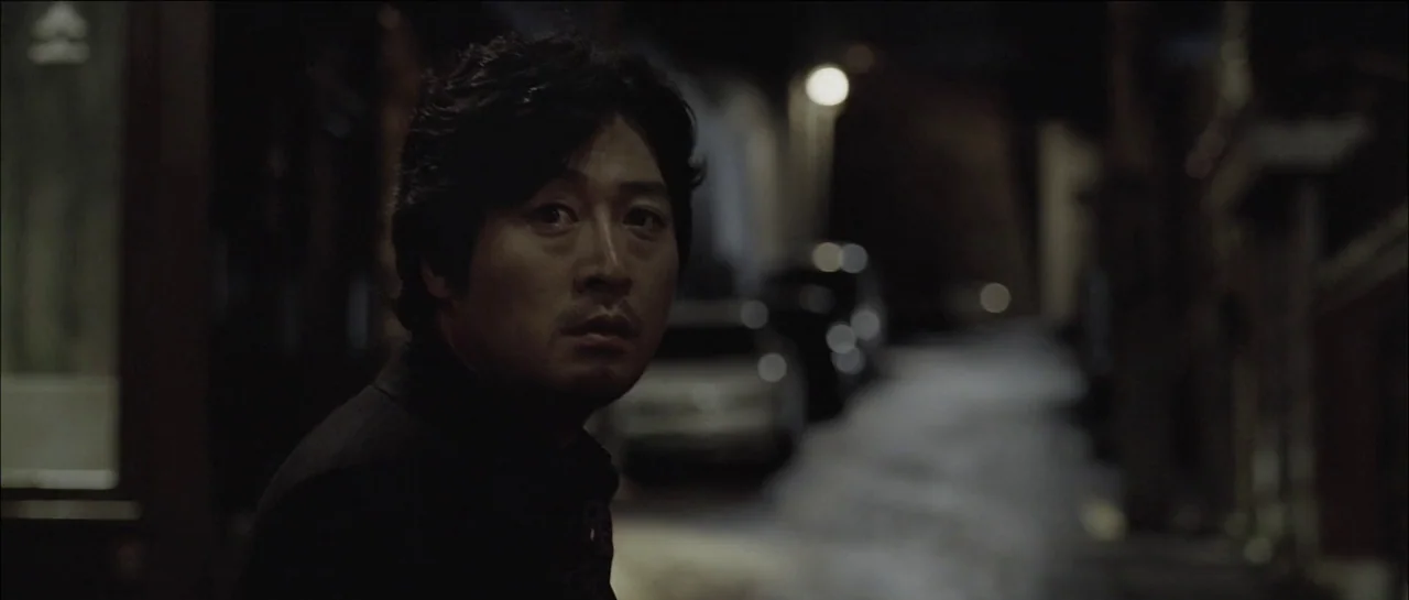 The Chaser (2008) movie still: medium — A dark, narrow urban alleyway is illuminated by a distant streetlamp at night. A middle-aged man stands in…