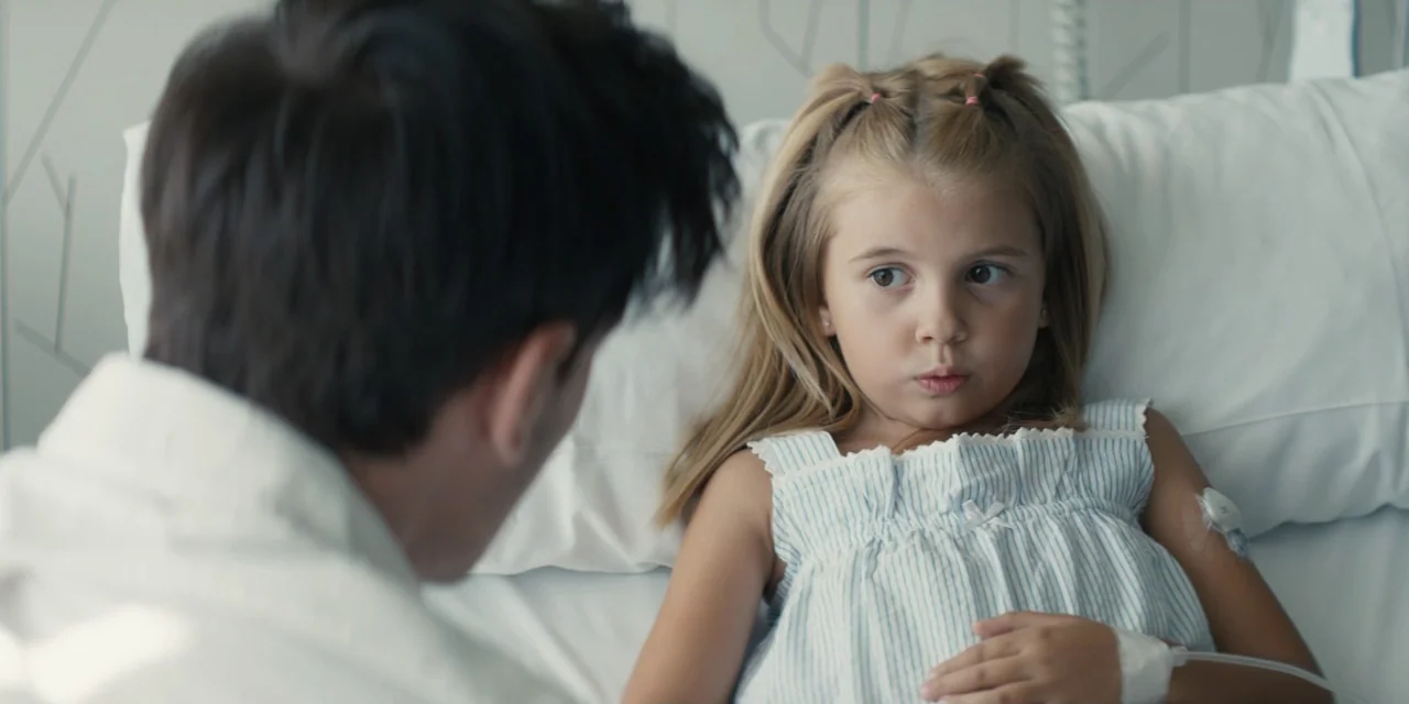 My Fault (2023) movie still: medium — A clean, bright hospital room features white pillows and medical equipment on the wall. A man sits beside the…
