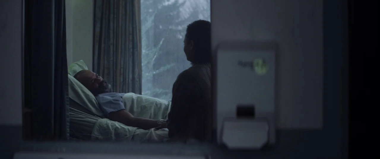 Hold The Dark (2018) movie still: medium — A hospital room with a patient bed positioned near a window with sheer curtains. A woman sits on a chair…