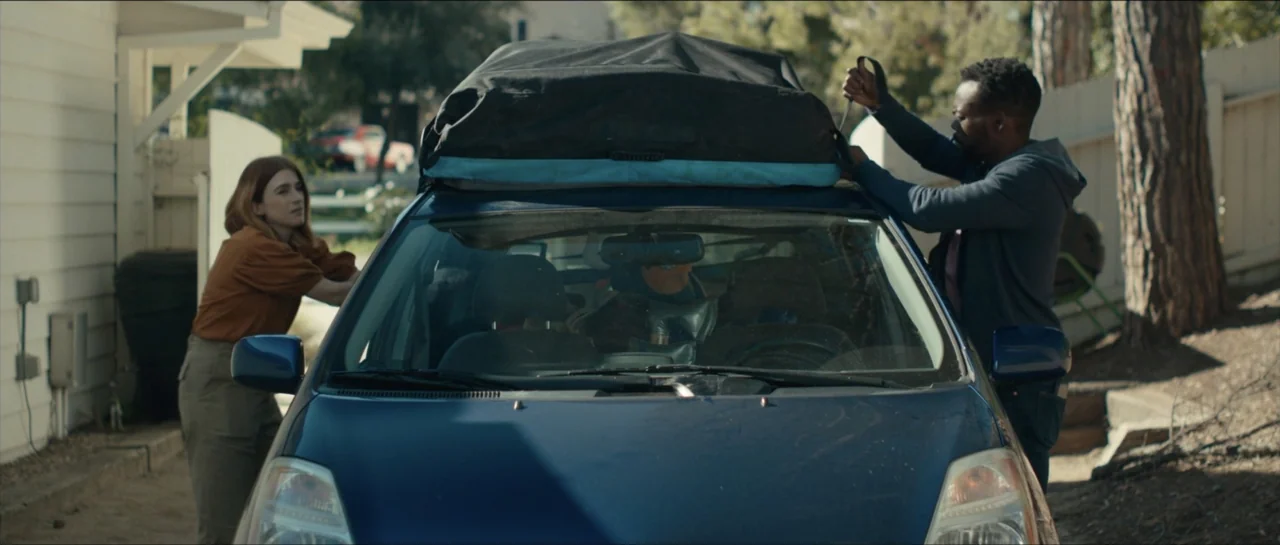We Broke Up (2021) movie still: medium — A suburban house exterior with a driveway holding a parked blue car. A man and woman work together to strap…