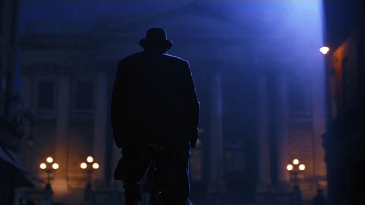 Michael Collins (1996) movie still: fullbody — A dark, classical building with pillars looms in the background of a night street. A silhouette of a…