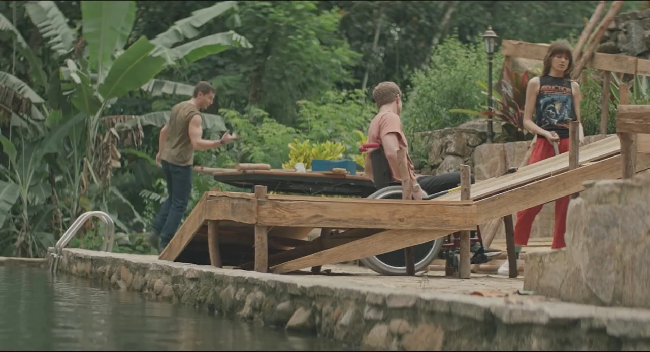 Proof of Love (2022) movie still: wide — A stone-edged pool terrace is surrounded by dense tropical foliage and banana trees. Three young adults…