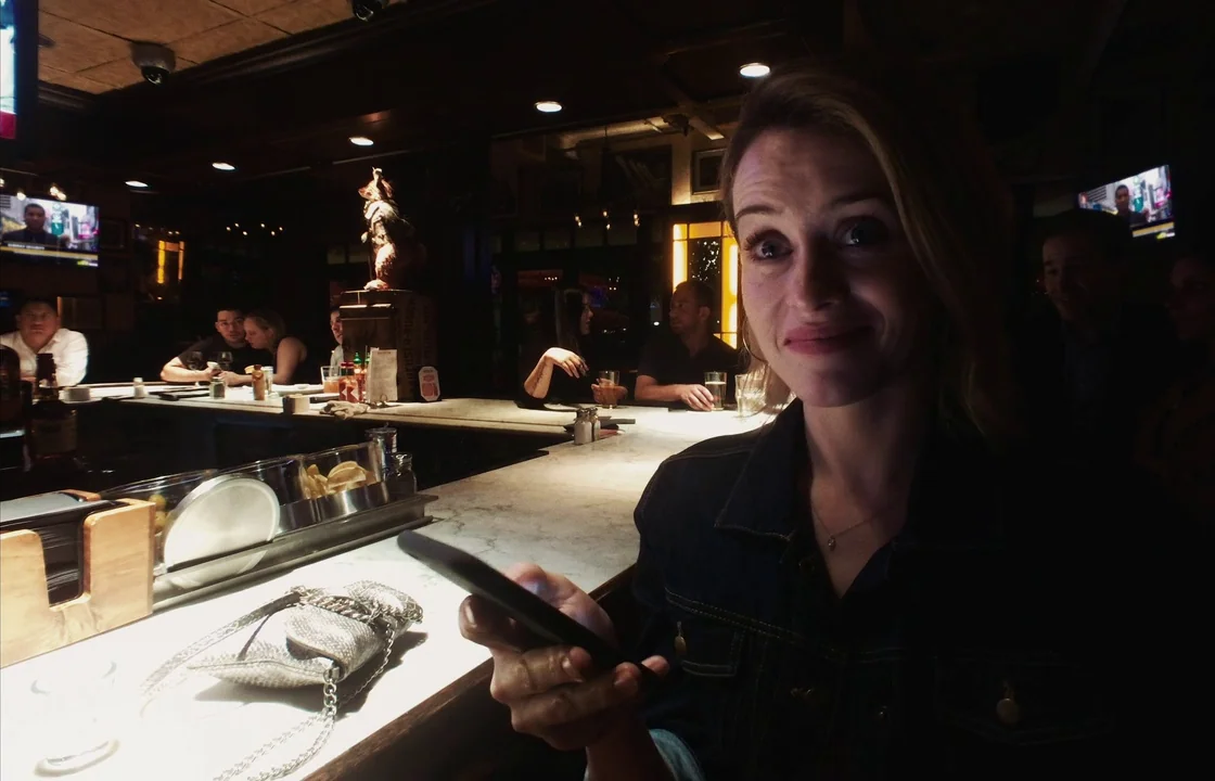 Unsane (2018) movie still: medium — A dimly lit bar interior features a long white marble top where patrons sit. A woman in a denim jacket holds a…