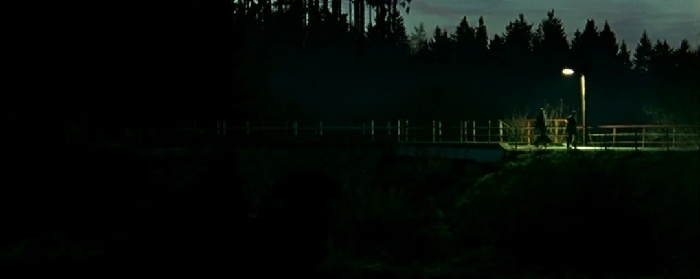 Calvaire (2004) movie still: wide — A dark, silhouetted bridge spans a hidden ravine under a faint night sky. Two figures walk slowly beneath the…